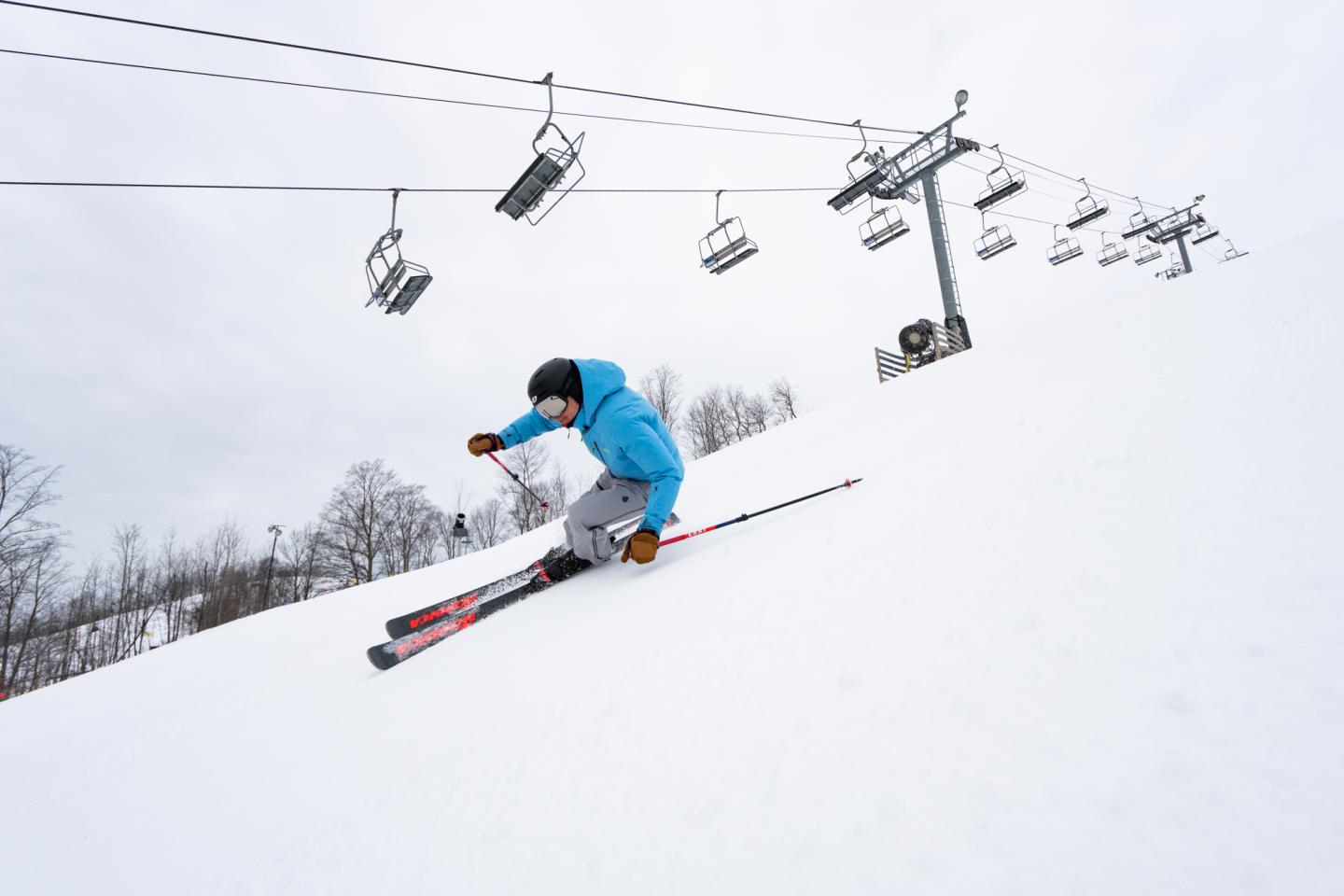 Skier in blue jacket descends snowy slope under a ski lift.