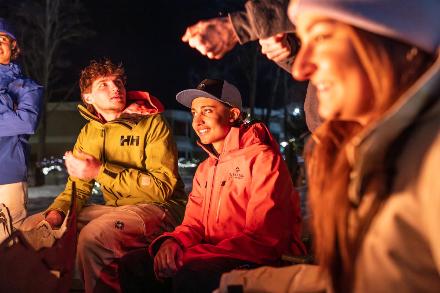Campfire gathering at night, people in warm jackets, smiling and talking.