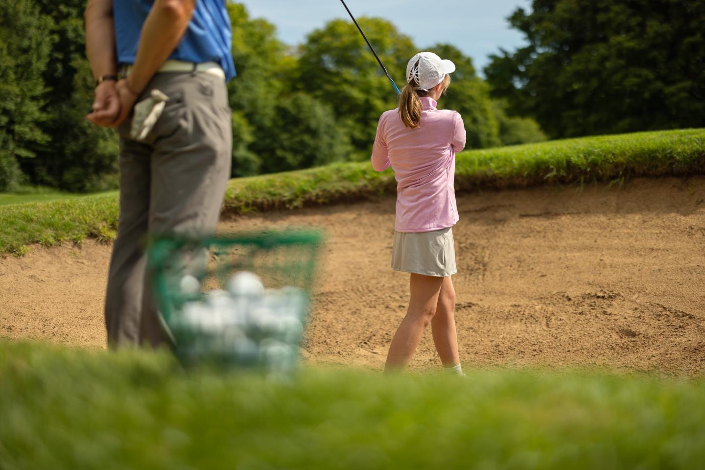 Golfer practicing swings near sand trap, with basket of balls on grass.