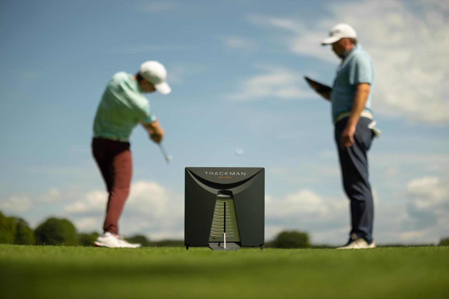Golfer swings club as another observes, with trophy in foreground under blue sky.