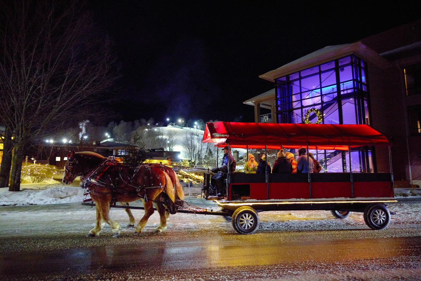 Horse-drawn carriage with passengers on a snowy night near a brightly lit building.