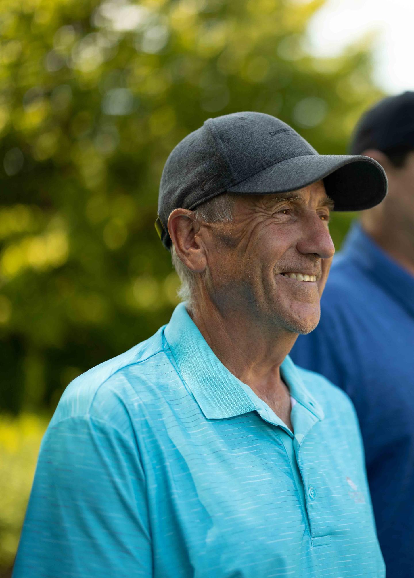 Smiling man in a blue shirt and cap, outdoors on a sunny day.