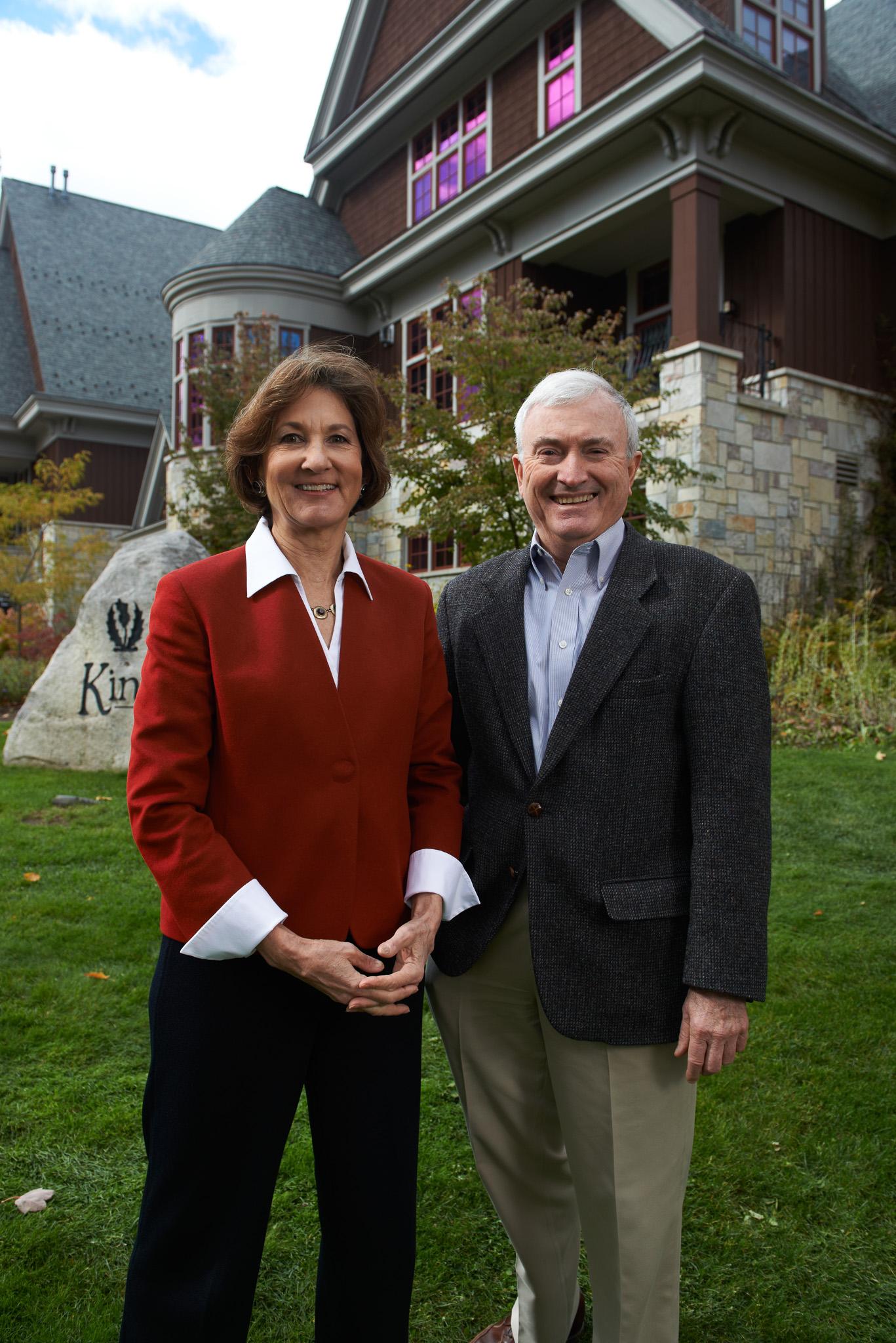 A man and woman smiling, standing in front of a large house.