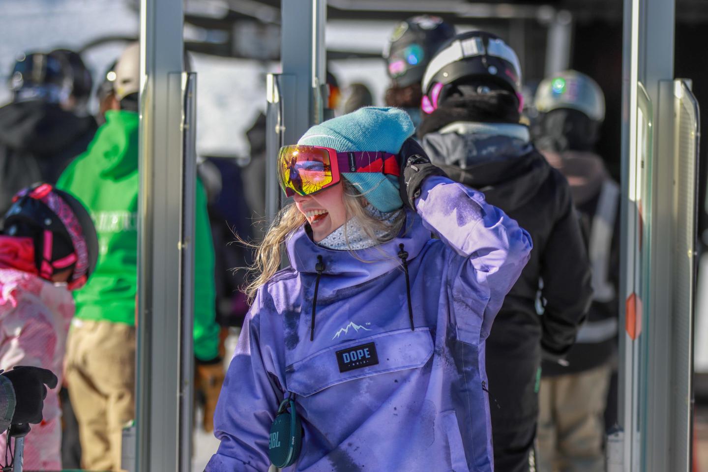 Snowboarder in purple jacket, adjusting goggles, surrounded by others at a ski lift.