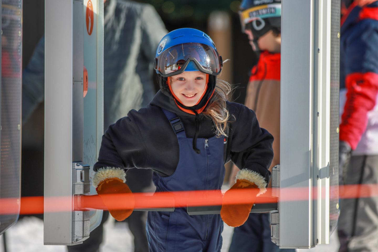 Child in ski gear and helmet smiling at a ski lift gate.