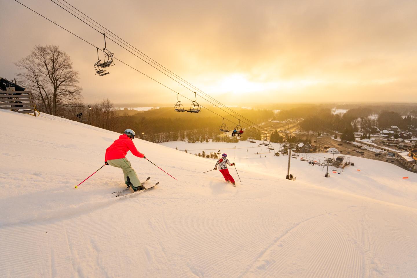 Skiers descend a snowy slope at sunset, with a warm glow and ski lift in the background.