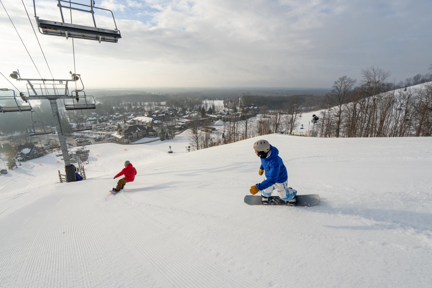 Snowboarders on a snowy slope, with a ski lift and town in the background.
