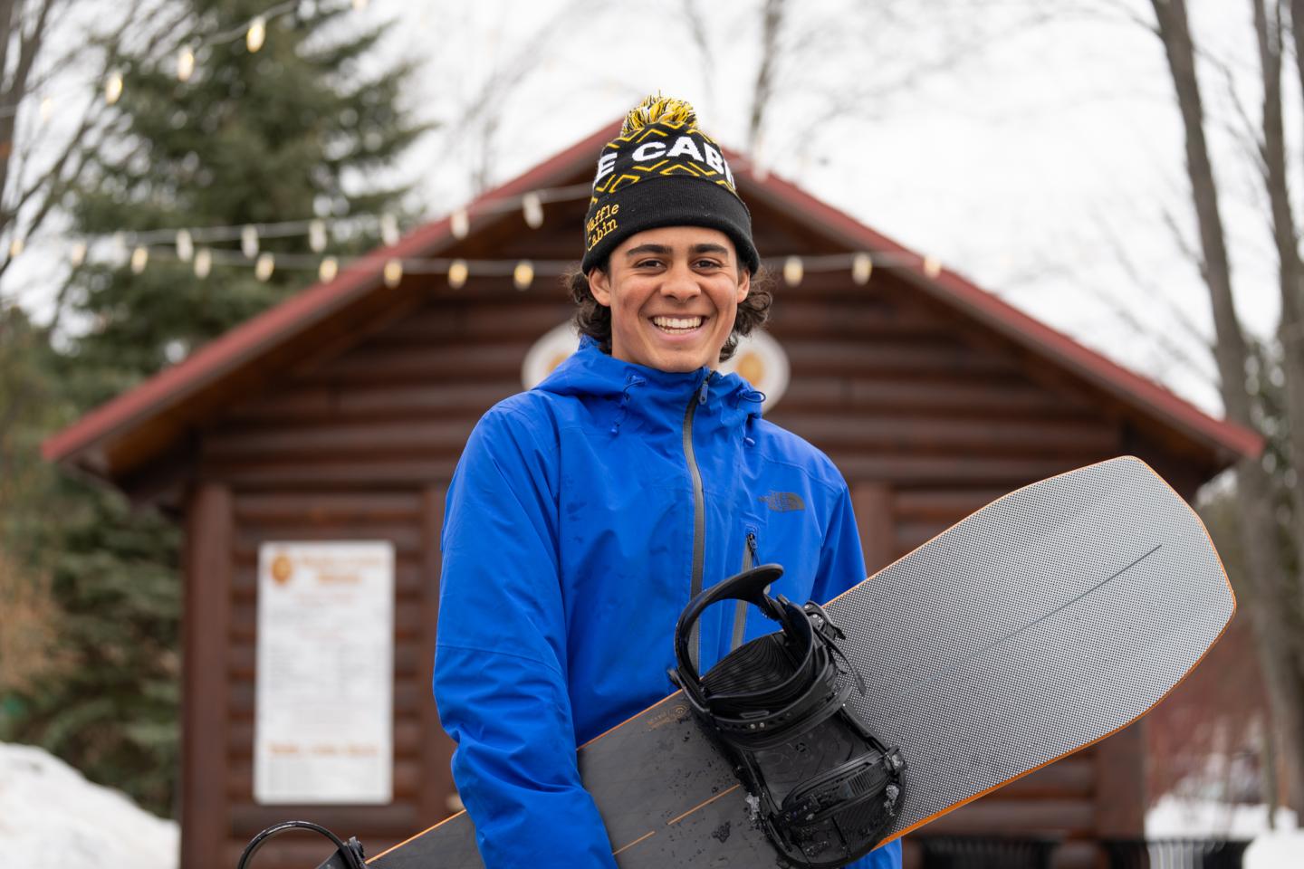 Smiling person holding a snowboard in front of a wooden cabin.
