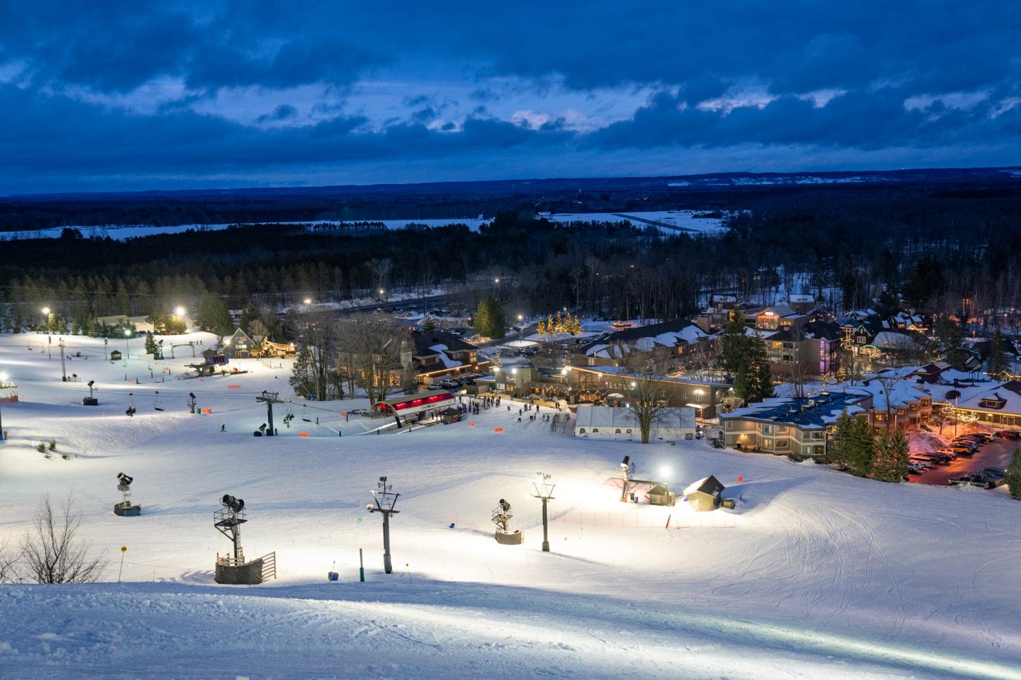 Snowy ski resort illuminated at dusk with visible ski lifts and buildings.