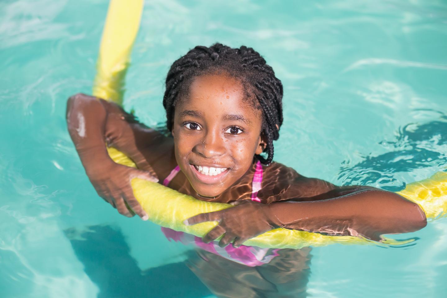 Young girl in a pool smiling, holding a yellow pool noodle.
