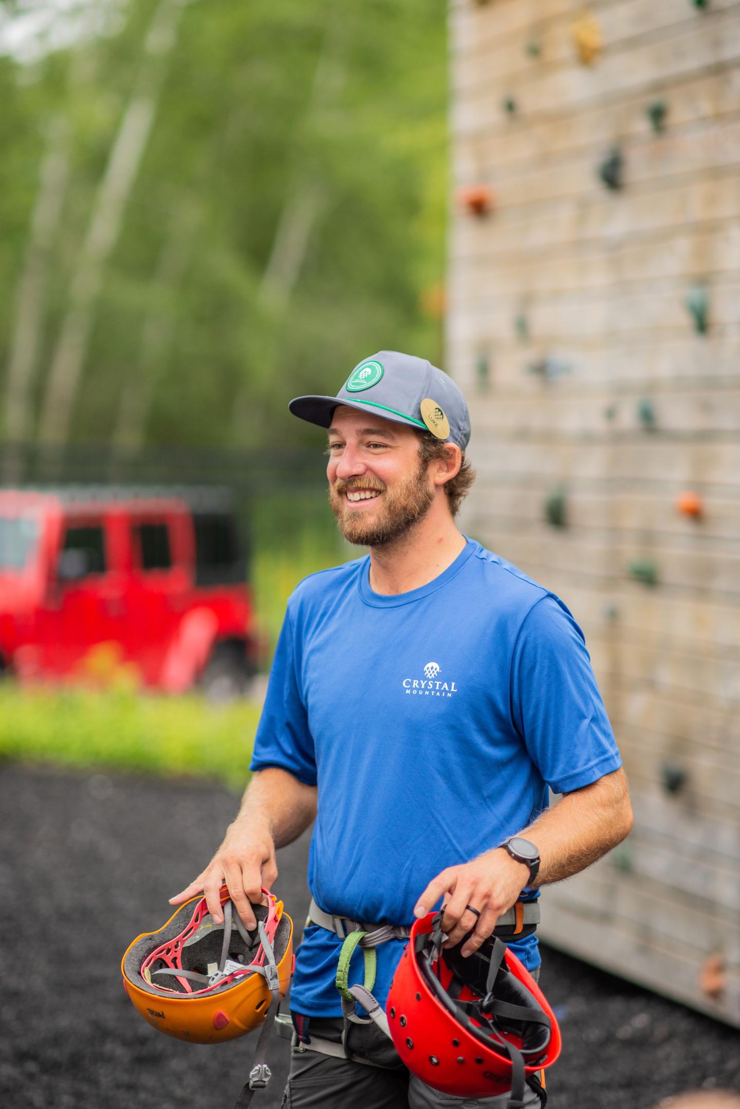 Man smiling outdoors, holding climbing helmets near rock wall.