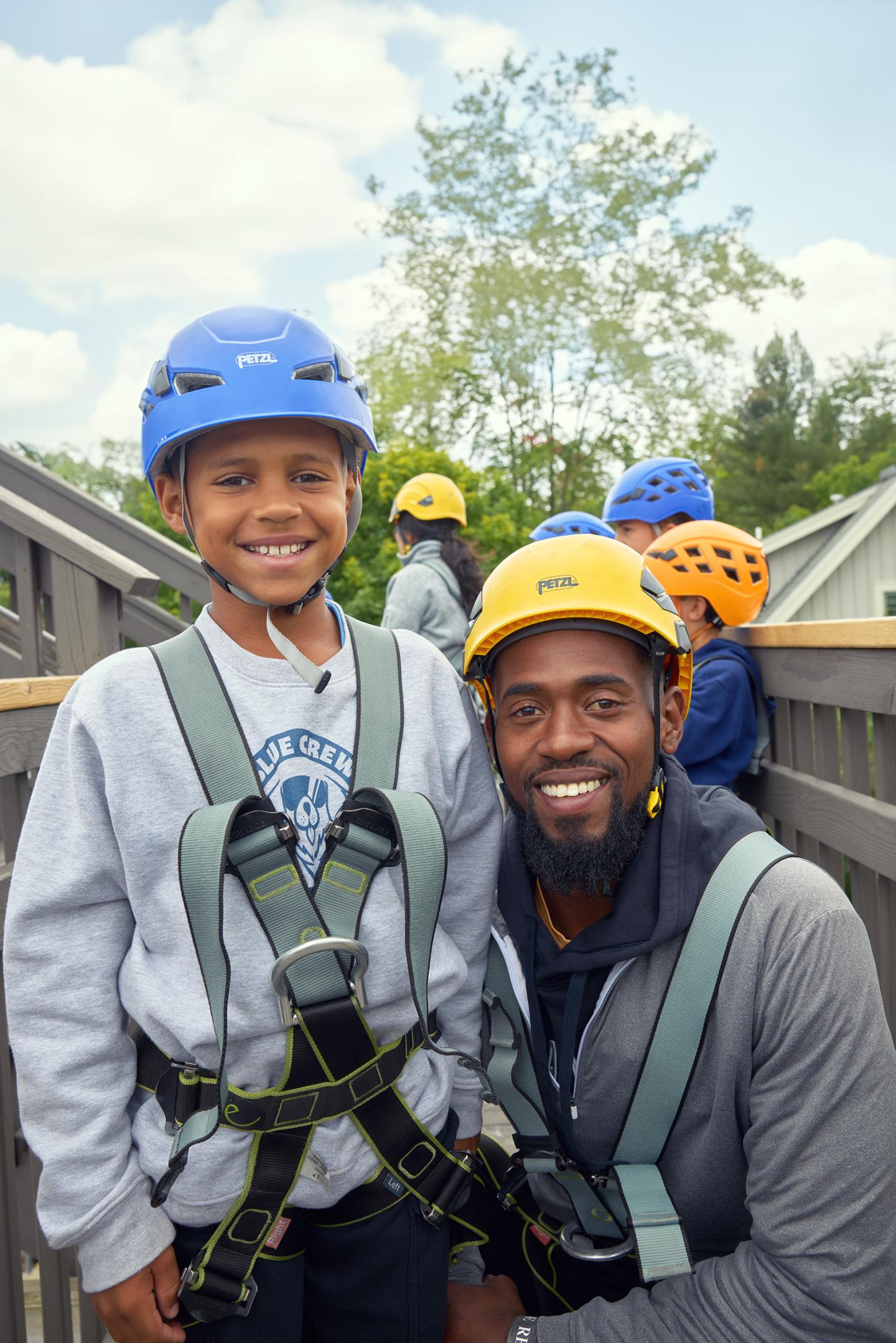 Father and son wearing helmets and harnesses, smiling in outdoor setting.