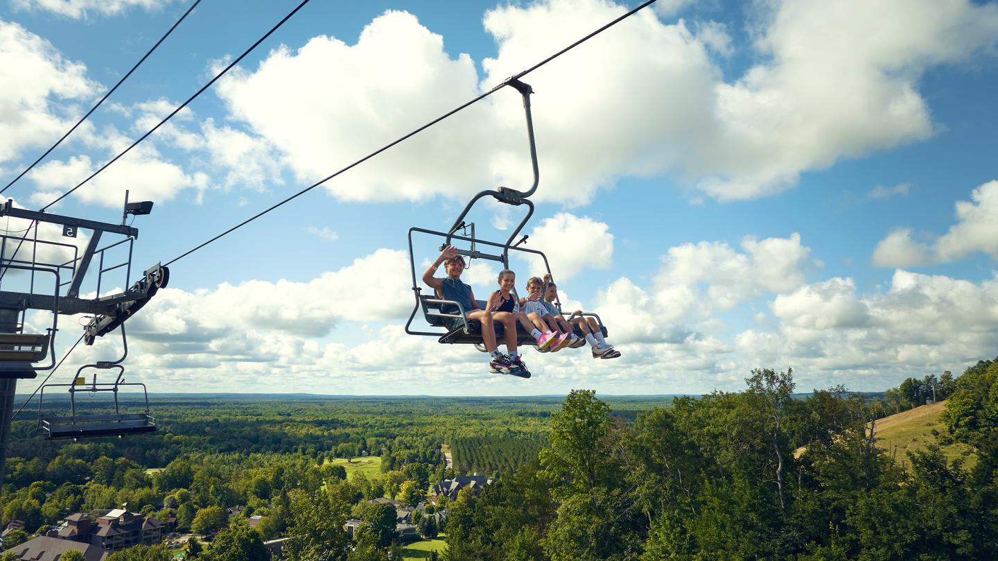 People on a chairlift with a scenic view of green hills and a blue sky with clouds.