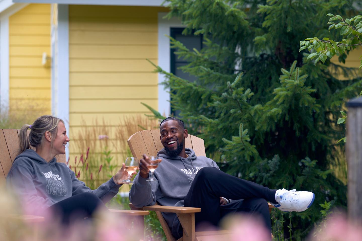 Two people smiling and toasting in Adirondack chairs by a yellow house and greenery.