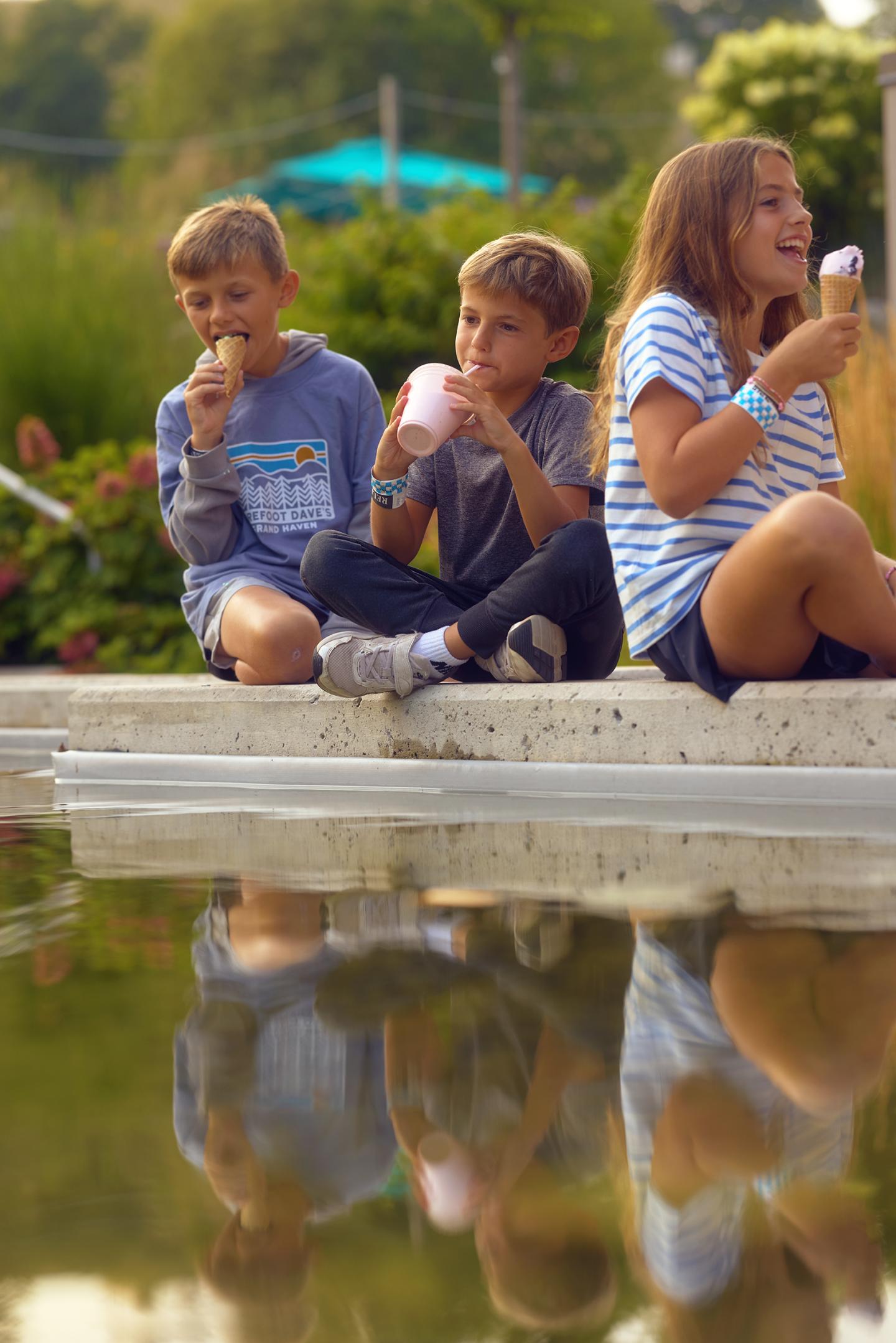 Three children enjoying ice cream by a pond with reflections on the water.