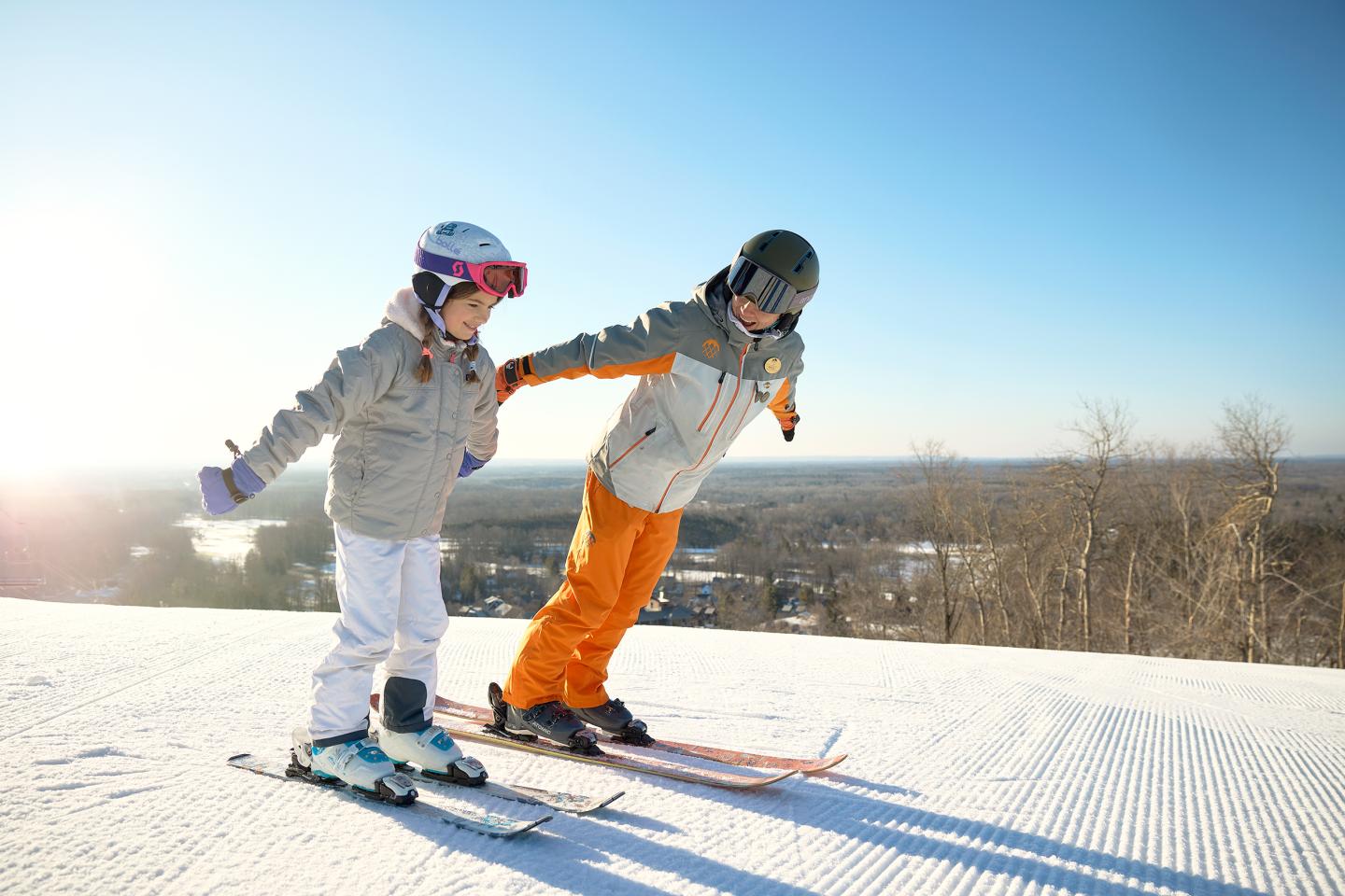 Two people skiing on a sunny slope, arms outstretched, with a winter landscape backdrop.