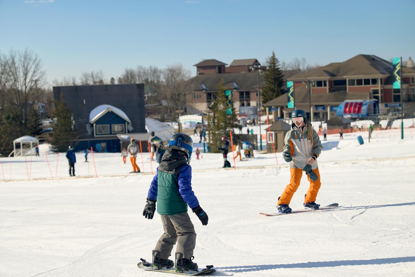 Two people snowboarding on a sunny ski slope near buildings.