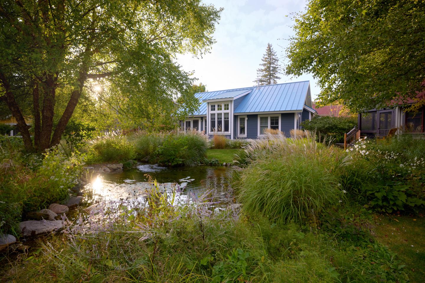 House with blue roof by a pond, surrounded by lush trees and greenery.