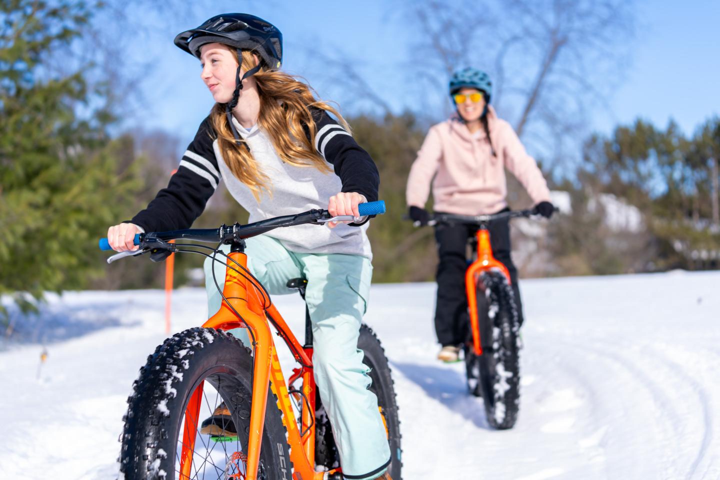 Two people biking on snow with orange fat bikes, wearing helmets in a sunny setting.