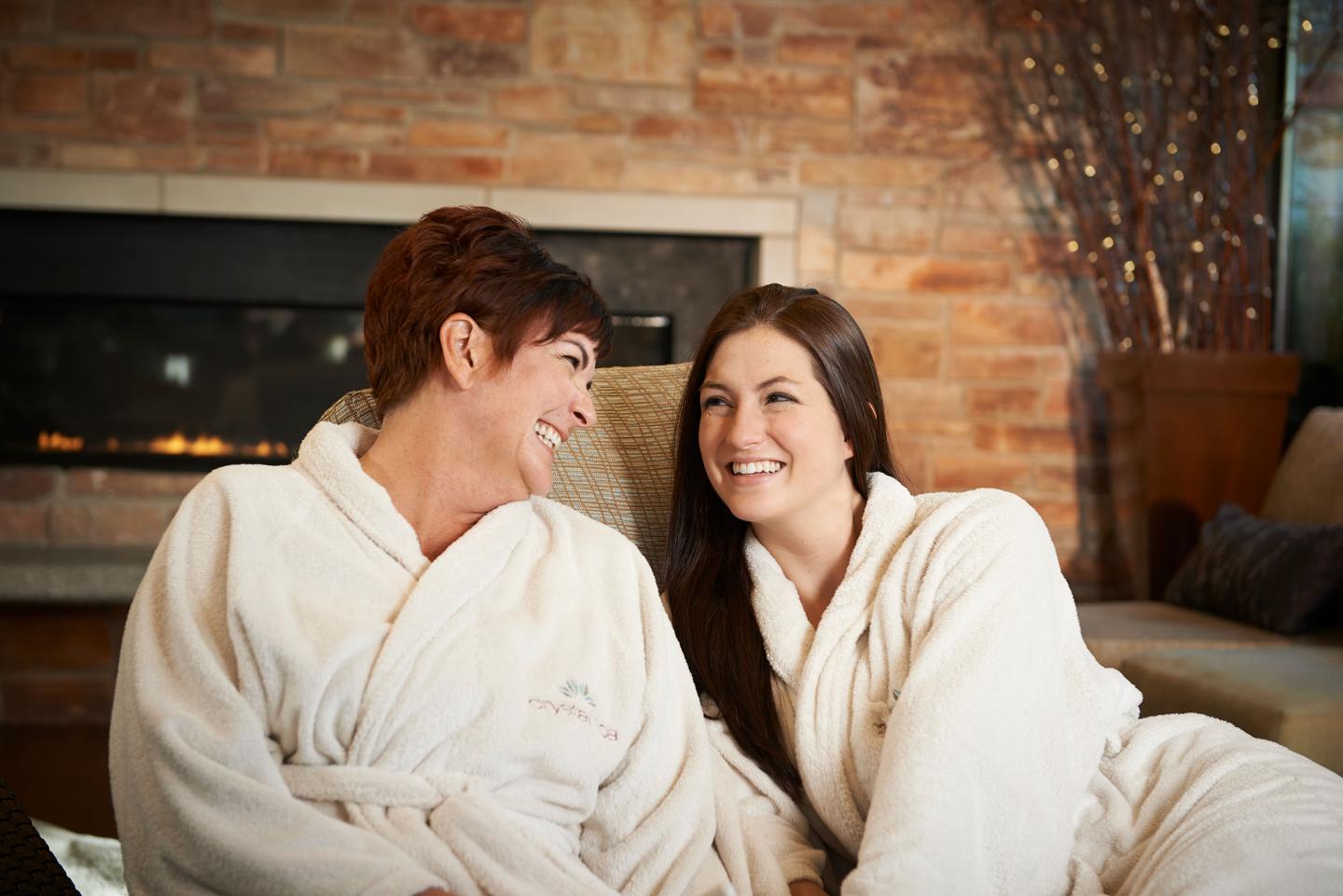 Two women in white robes smiling near a cozy fireplace.