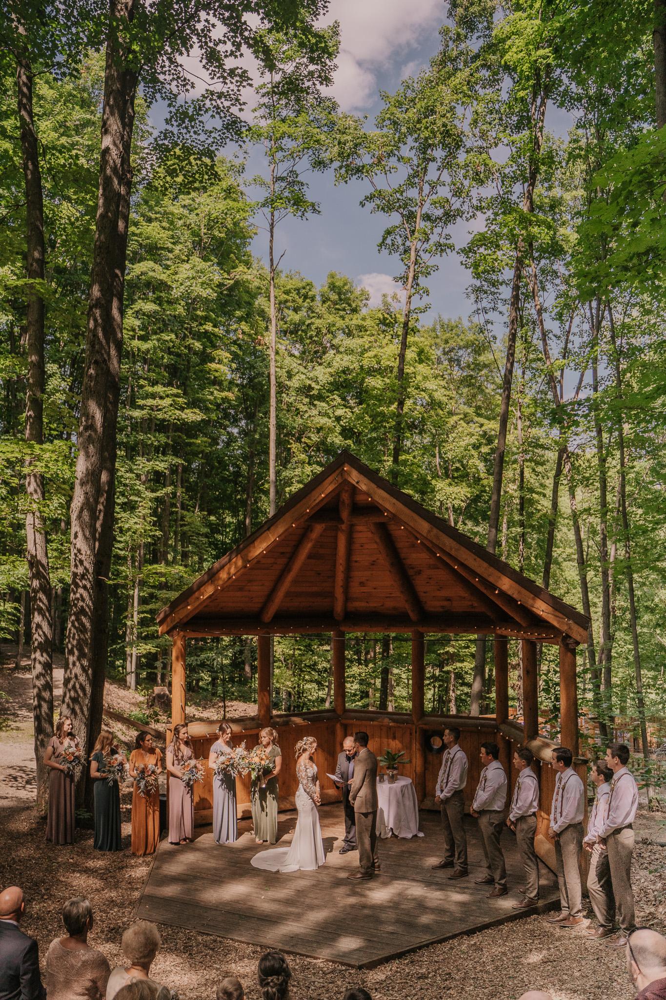 Forest wedding ceremony under wooden pavilion, guests seated, trees surrounding.