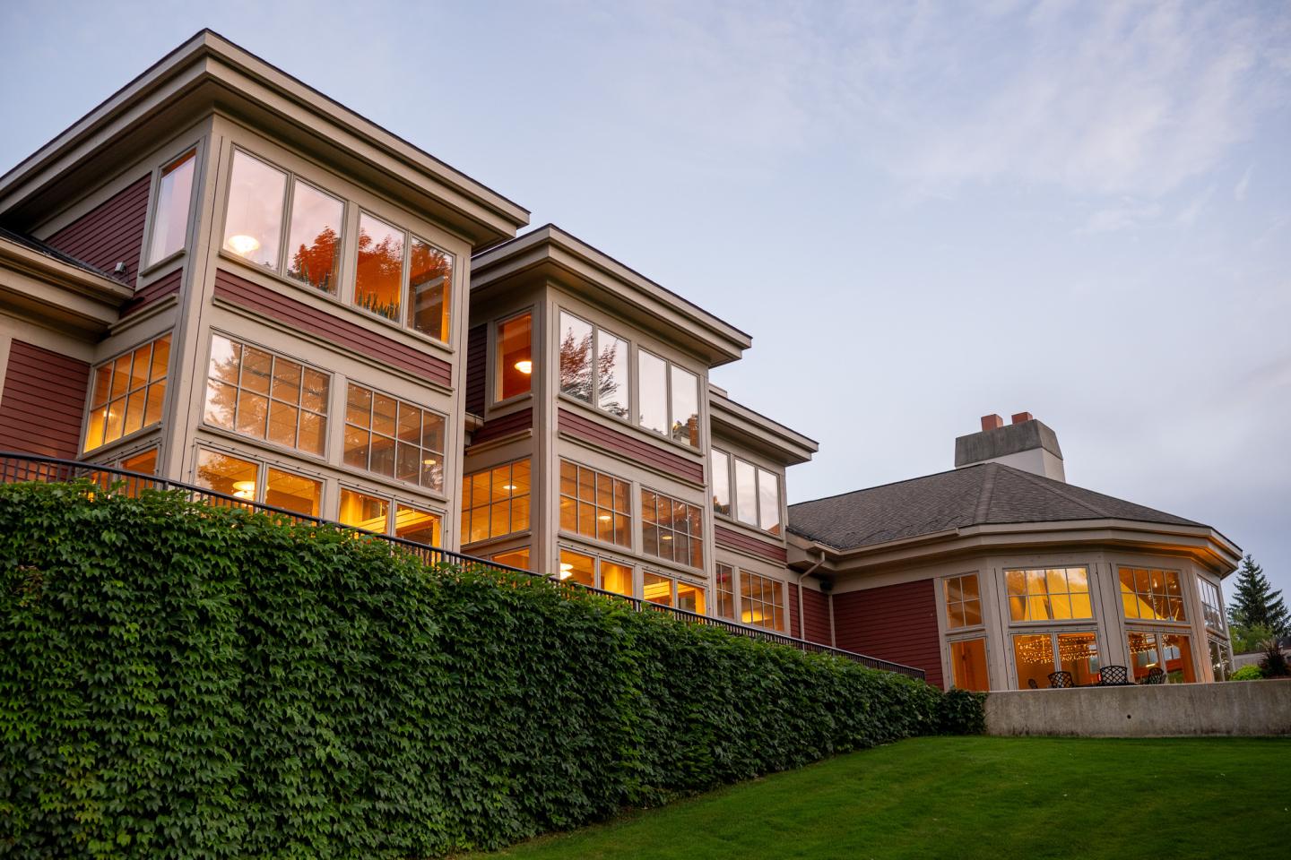 Large house with warm glowing windows and hedge, under a cloudy sky.