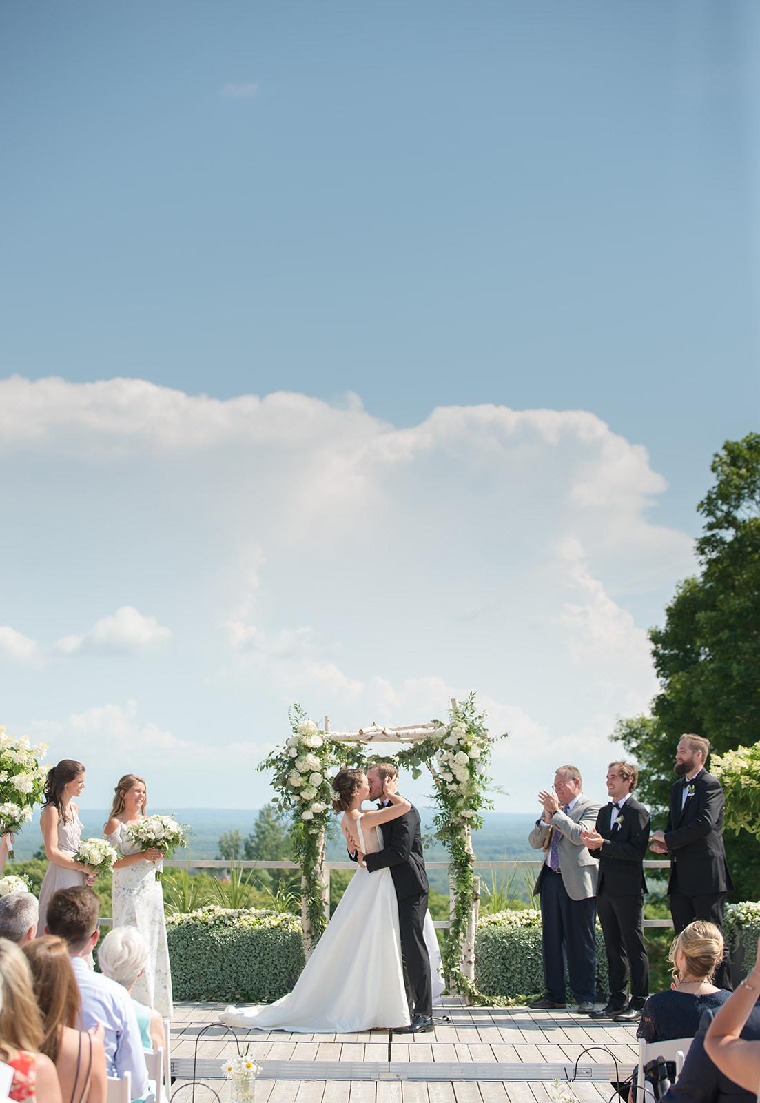 Bride and groom kiss under floral arch at outdoor wedding ceremony.