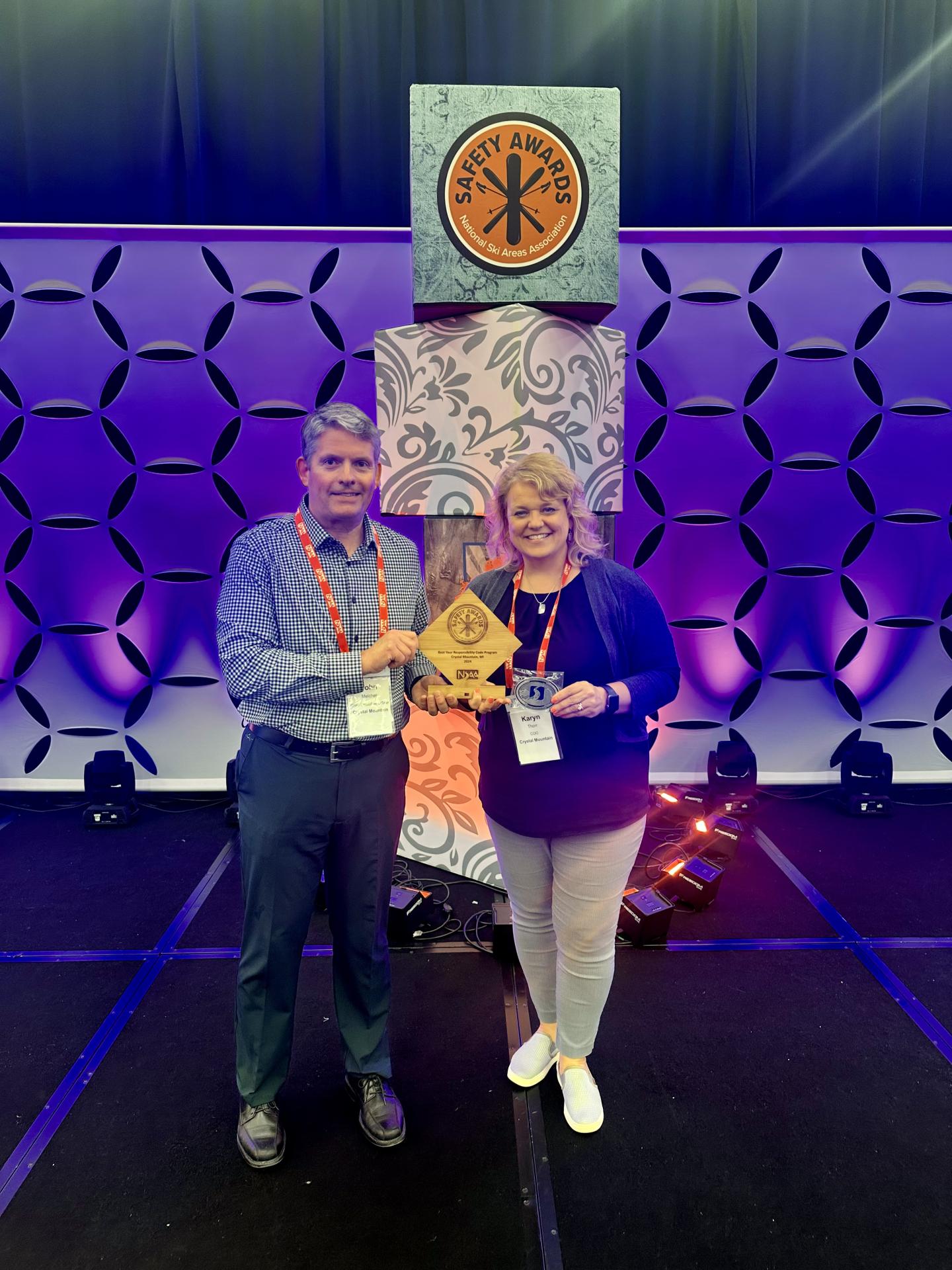 Two people smiling, holding an award on a stage with a geometric backdrop.