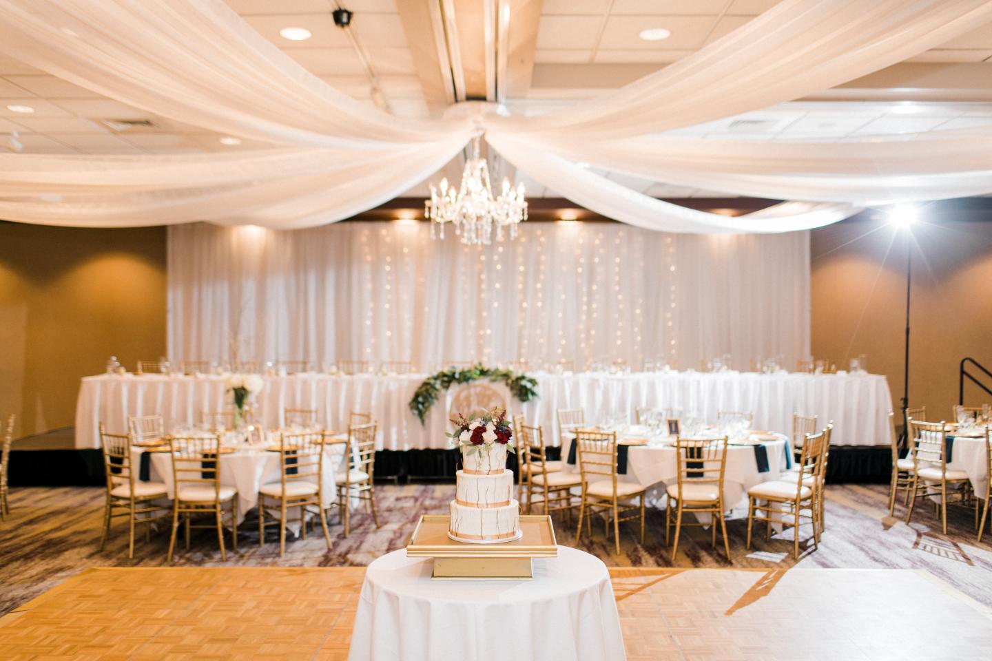 Elegant wedding reception with white drapes, chandelier, and floral-decorated cake table.