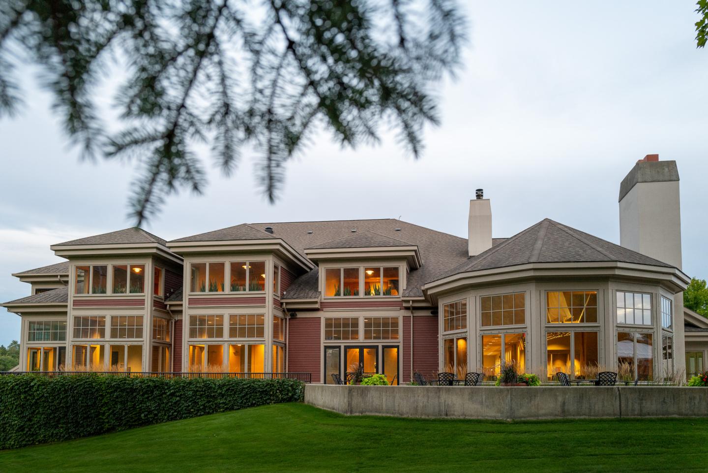Large house with illuminated windows, surrounded by green lawn and trees.