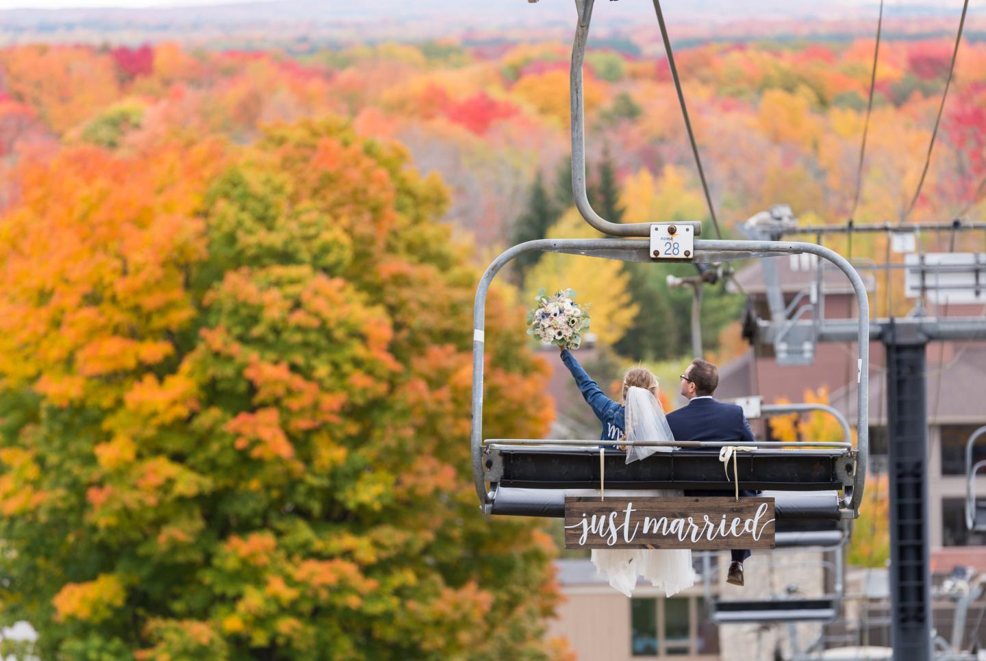 Newlyweds on a ski lift with autumn trees in the background.