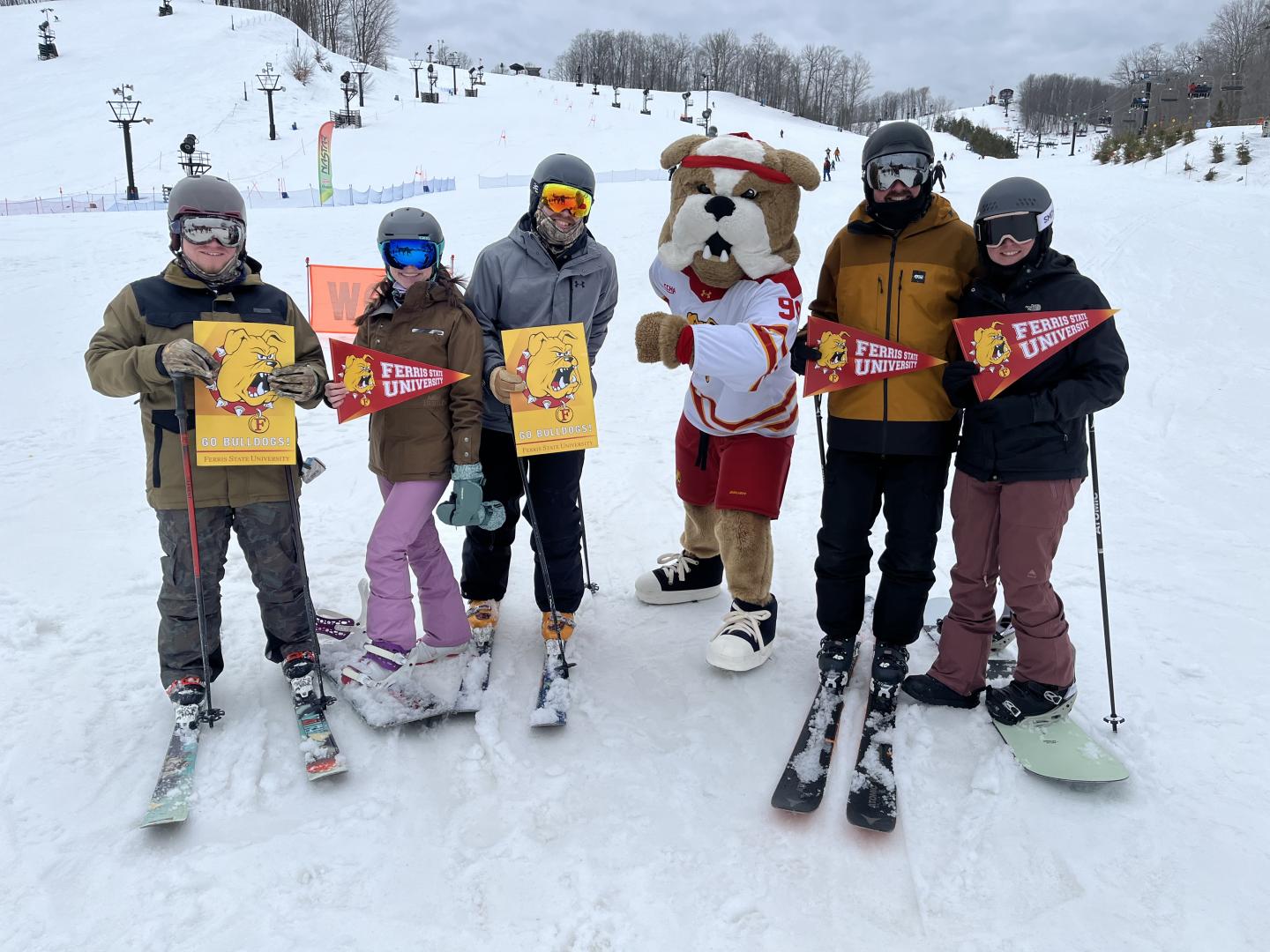 Skiers pose with a dog mascot on a snowy slope, holding signs and wearing winter gear.
