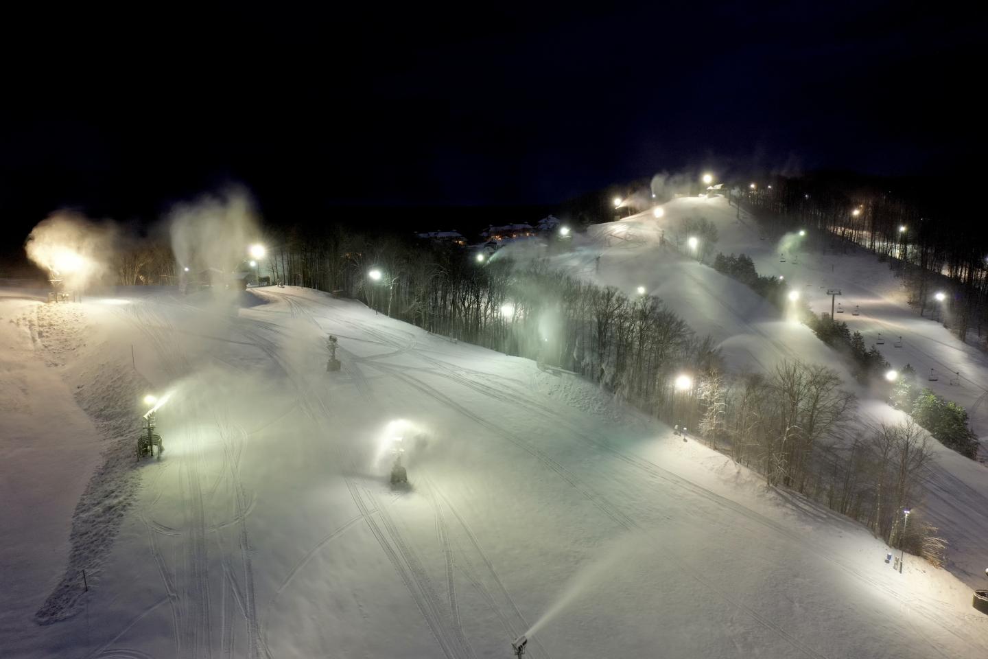 Snowy ski slopes lit at night with snow machines active.