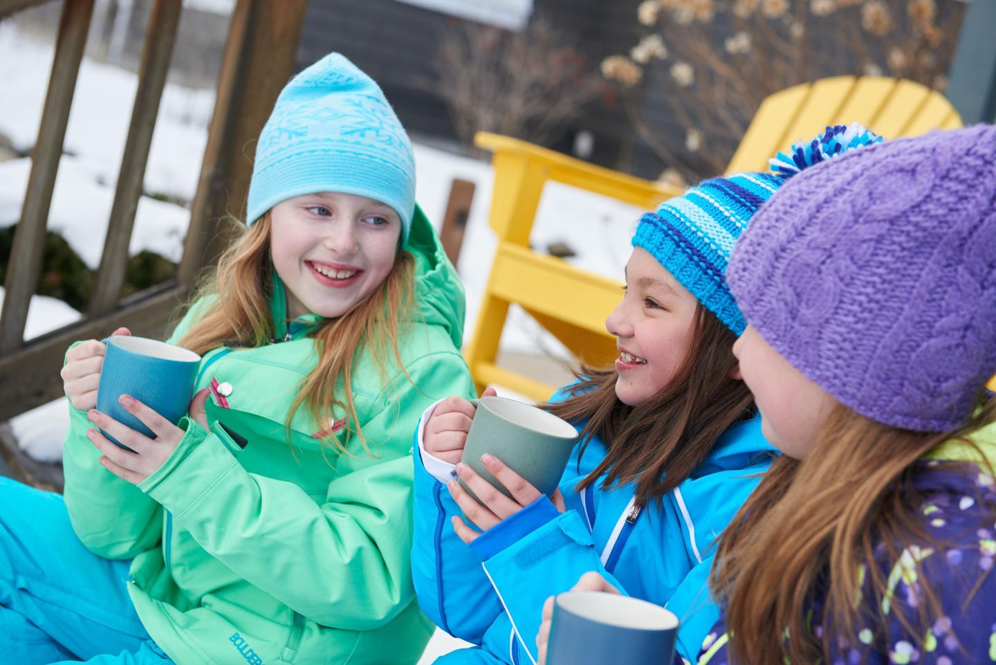 Kids in winter clothes, smiling, drinking from mugs outside.