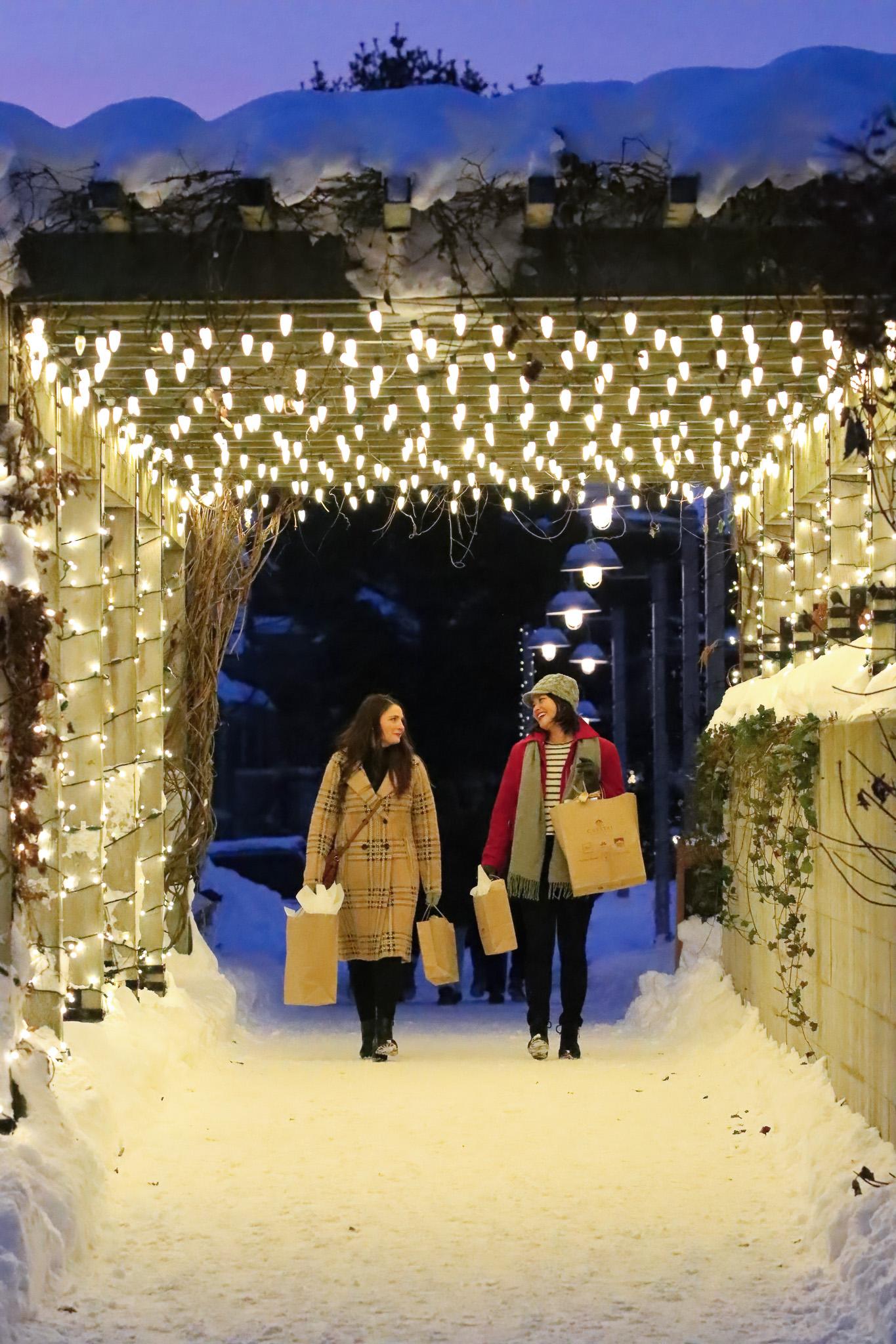 Two people walking under festive lights in a snowy pathway, carrying shopping bags.