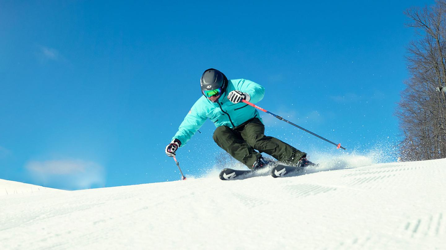 Skier in turquoise jacket on snowy slope under clear blue sky.