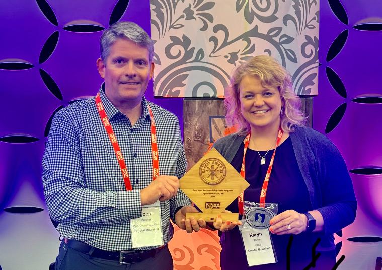 Two people smiling while holding wooden awards at an event with a patterned backdrop.