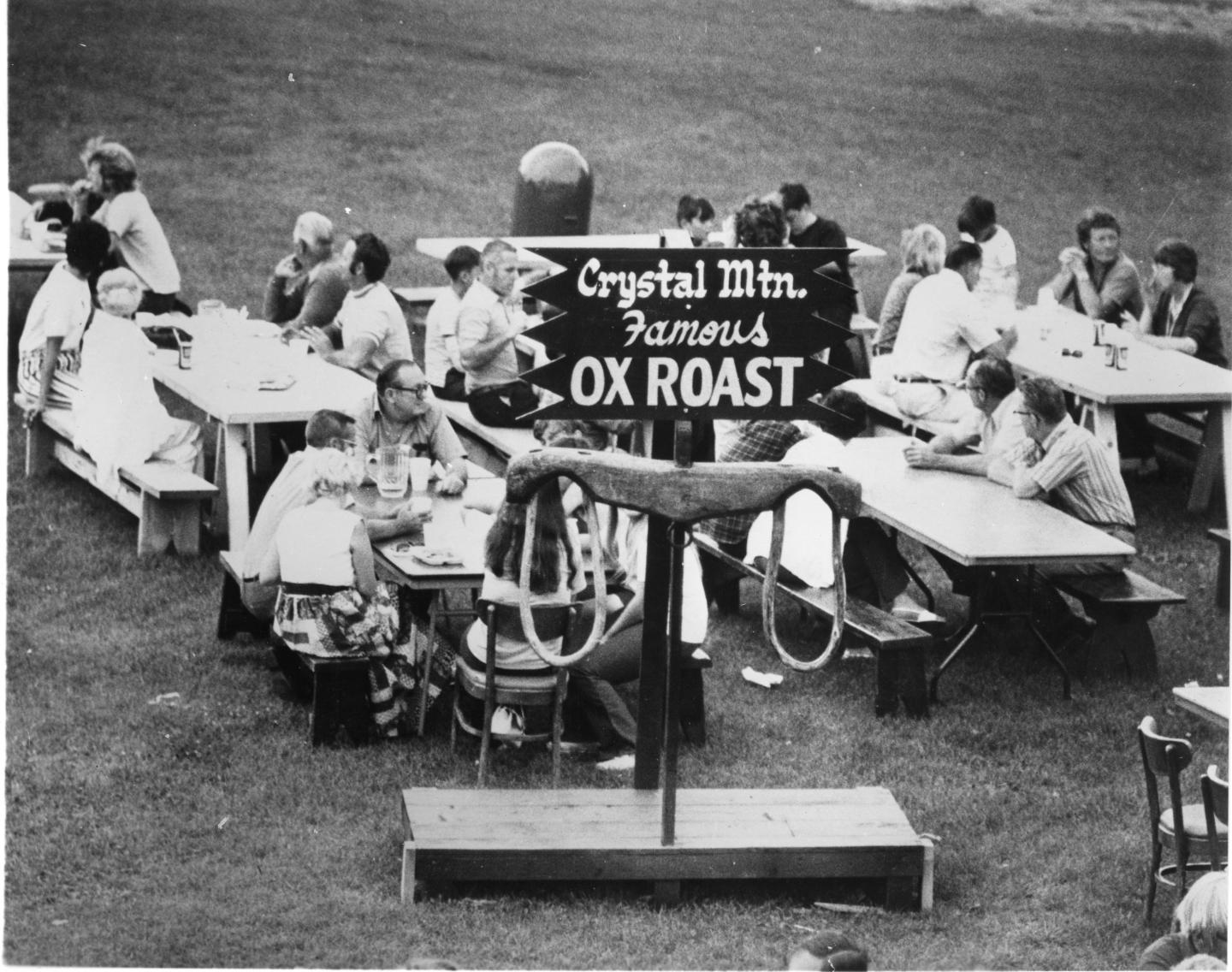 People seated at picnic tables outside, attending an ox roast event.