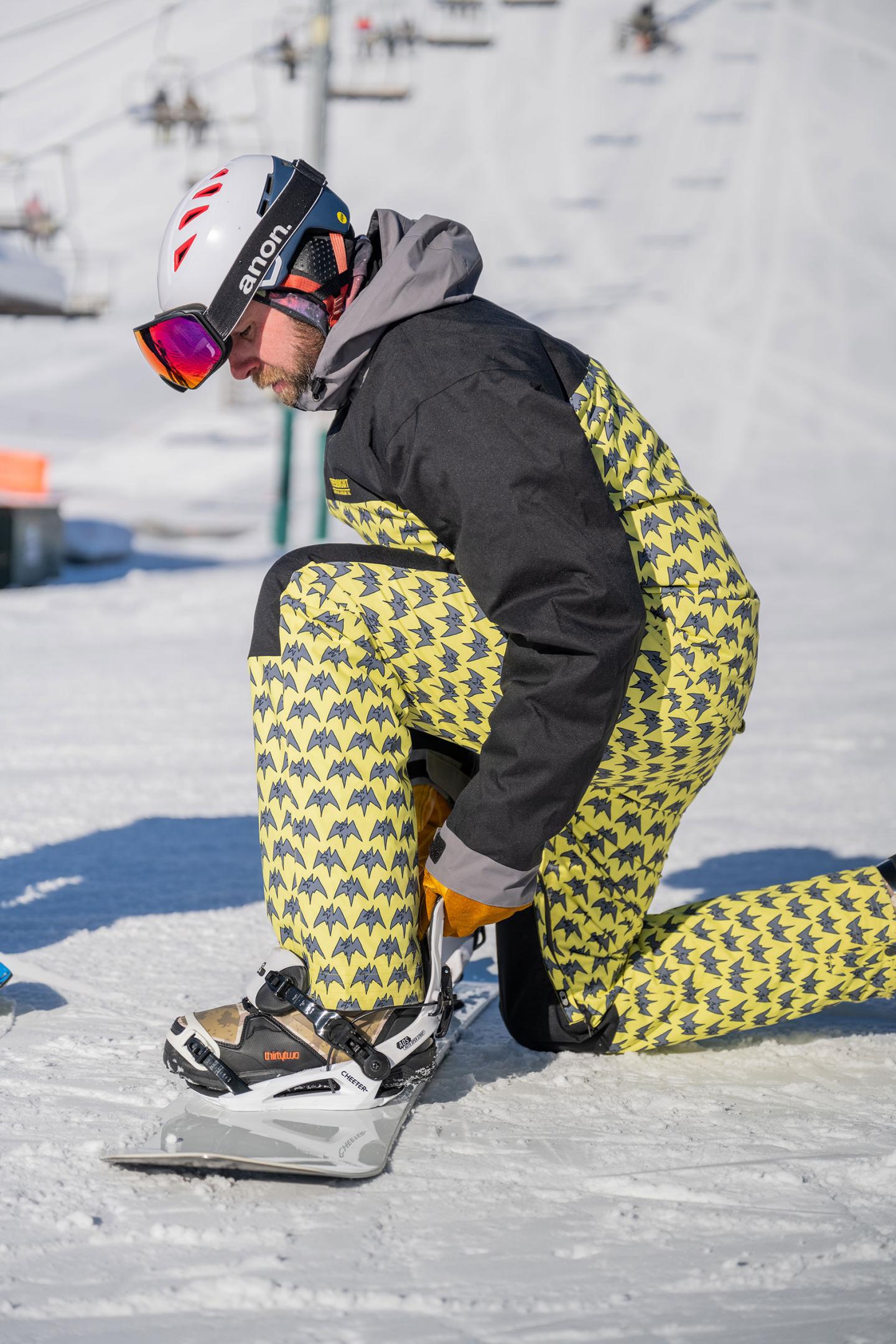 Snowboarder adjusting binding on a snowy slope, wearing yellow patterned pants.