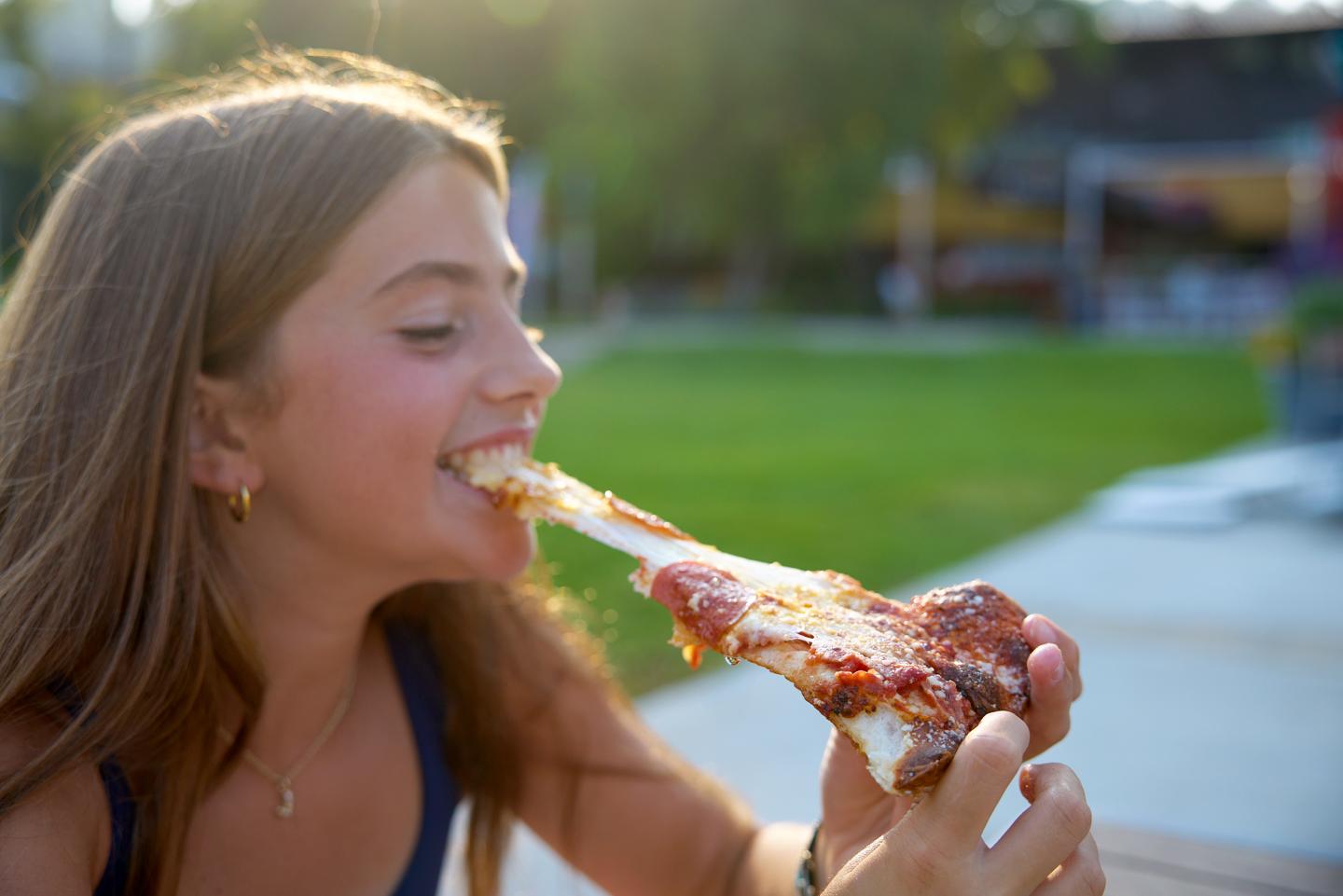 Girl enjoying a slice of pizza outdoors with cheese stretching.