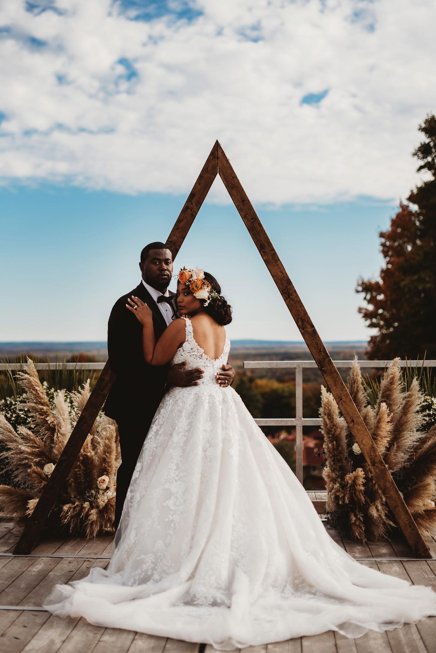 Bride and groom embrace under wooden triangle arch, against a scenic outdoor backdrop.