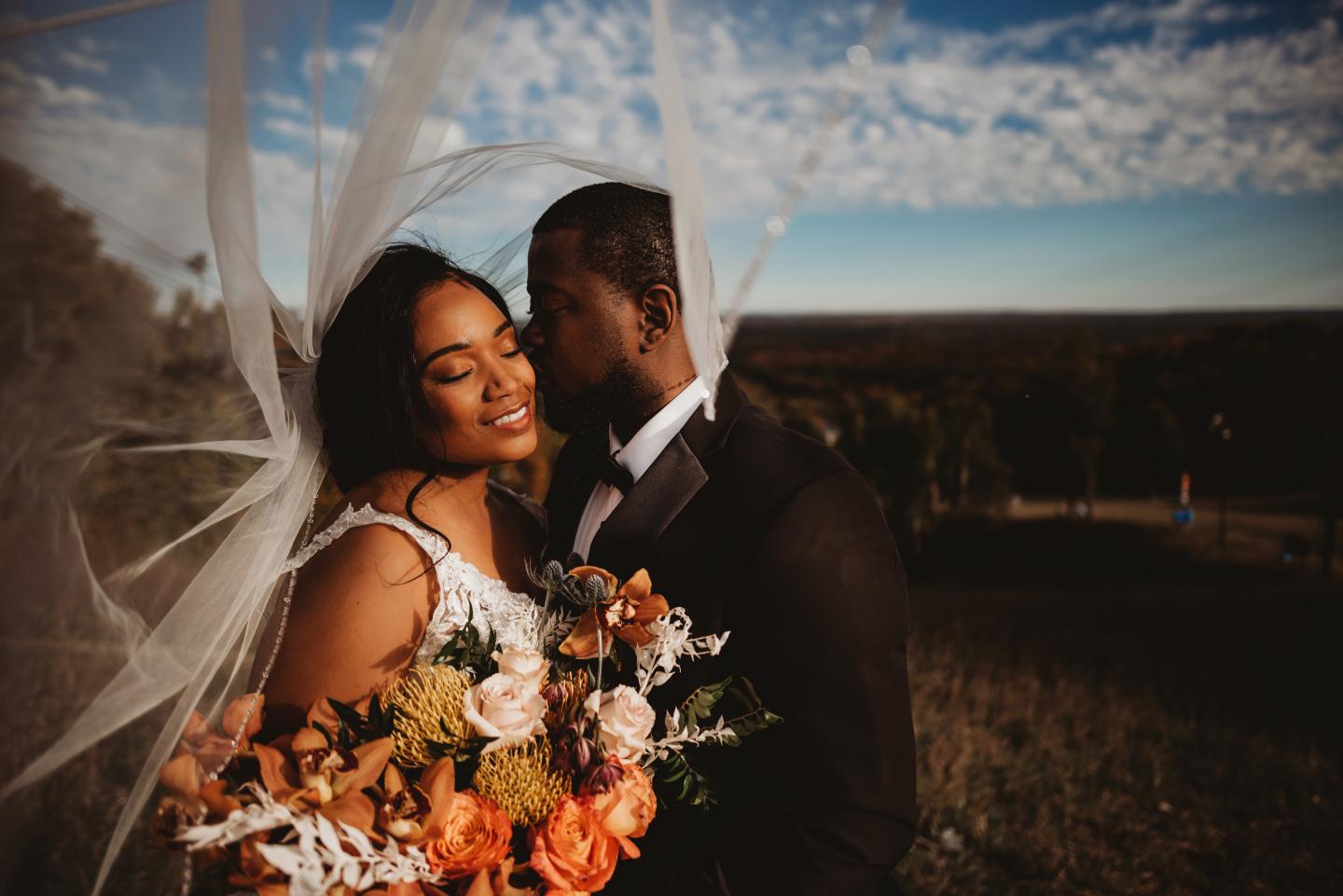 Bride and groom embrace with bouquet, blue sky in background.