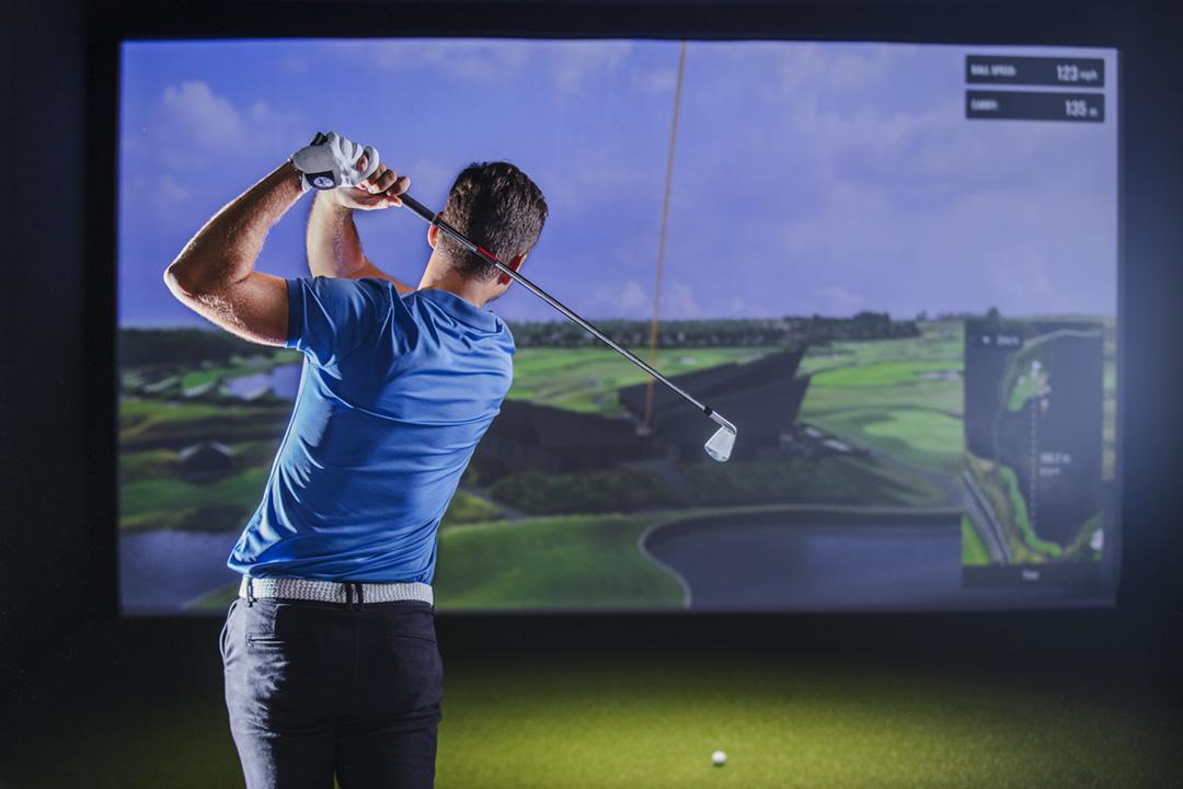 Golfer swinging in indoor simulator room with course projection.