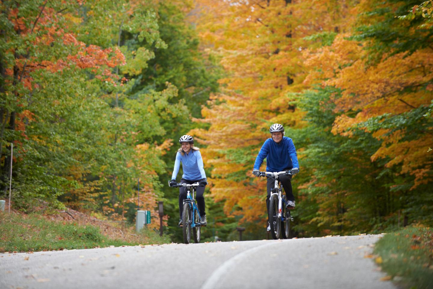 Two people biking on a tree-lined road with vibrant autumn foliage.