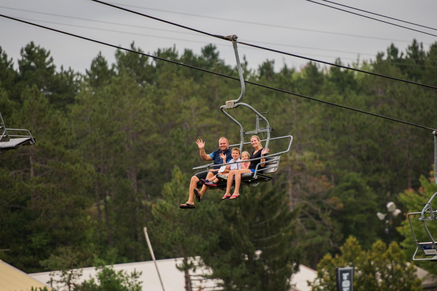 Family riding a ski lift over green trees, one person waving.