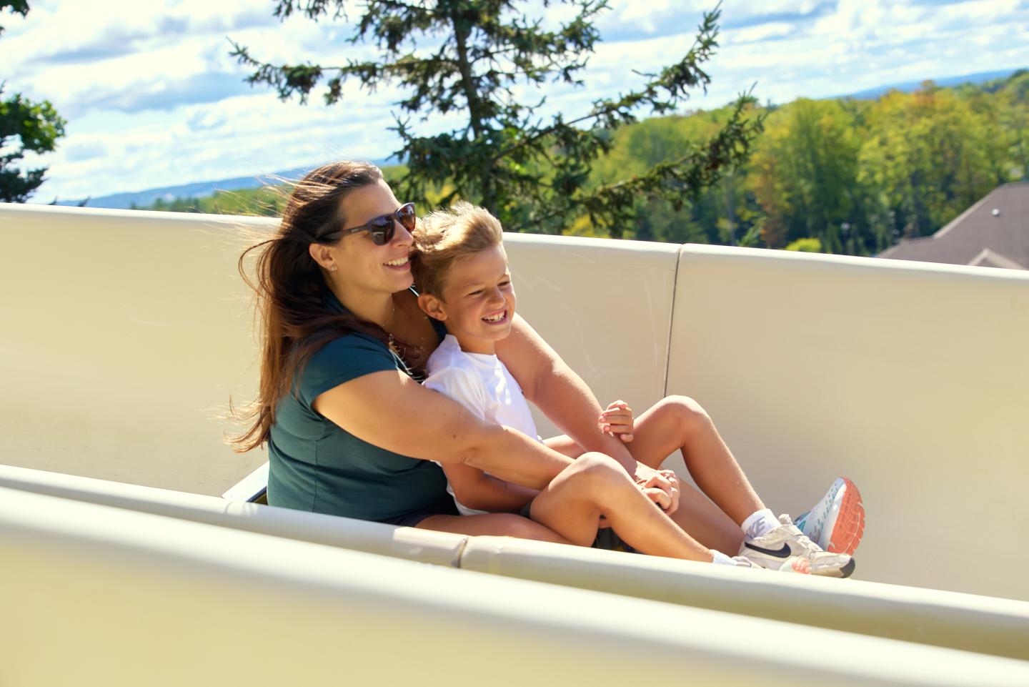 Mother and child laughing on a slide, sunny day, trees in background.