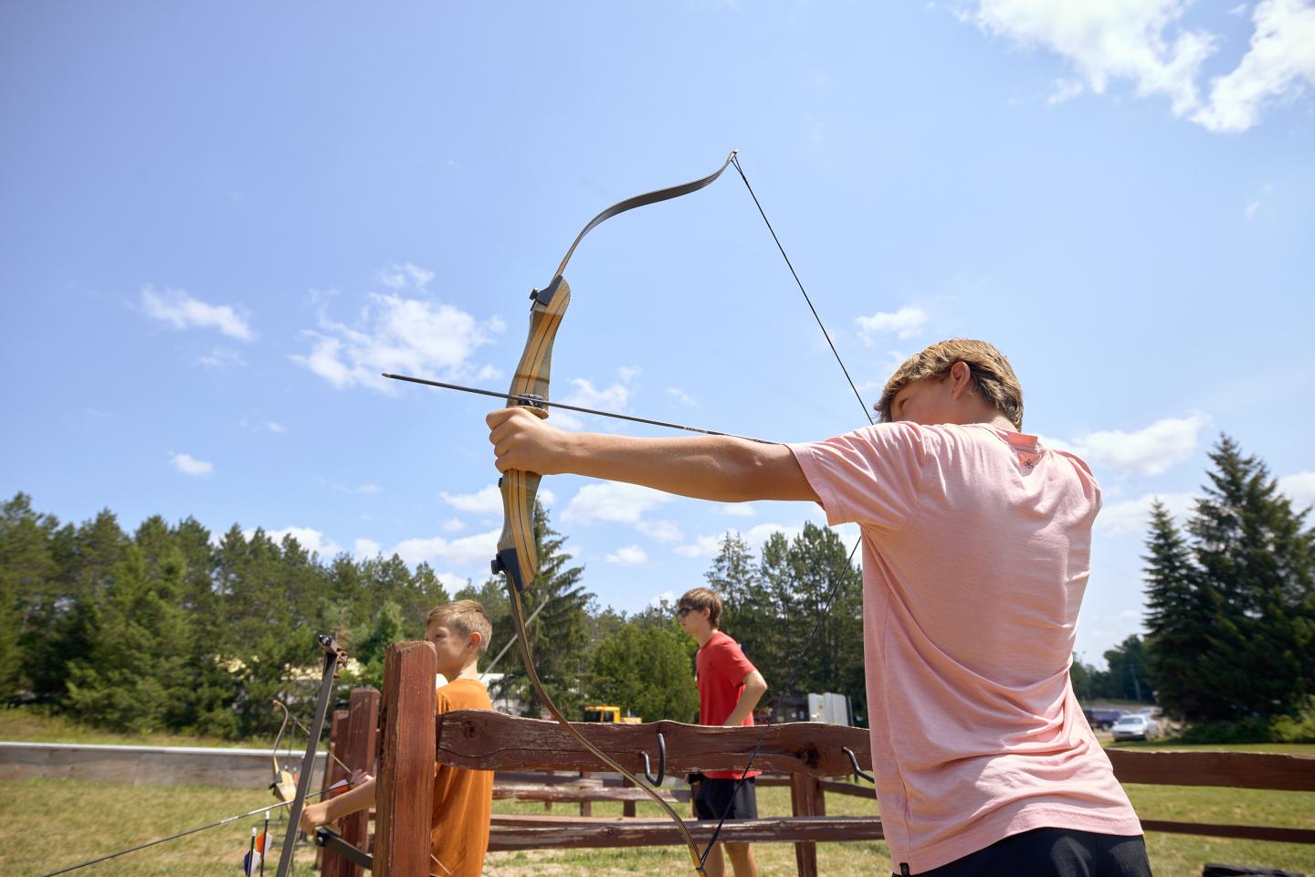Boy aiming a bow and arrow outdoors under a clear sky.