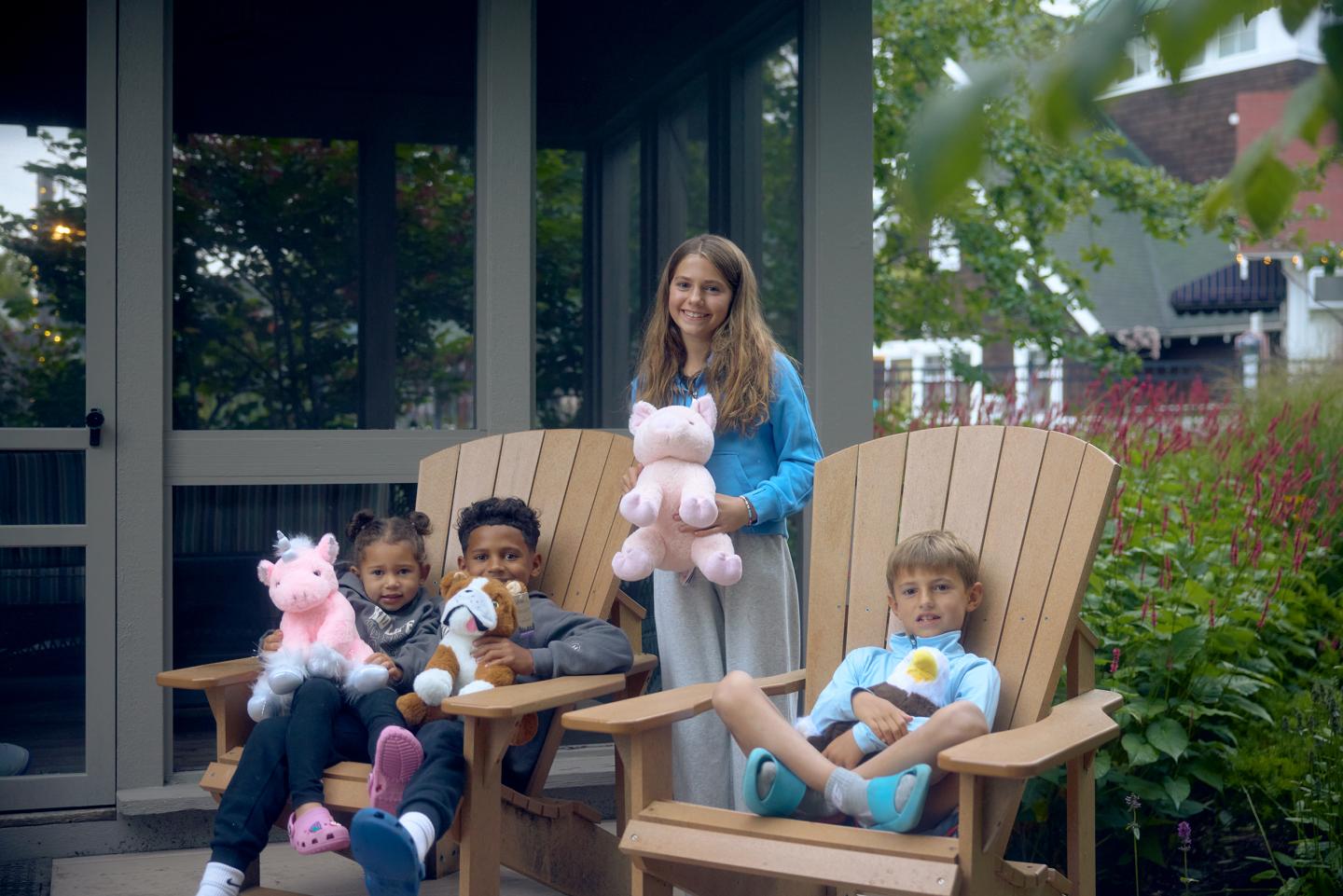 Children sitting on wooden chairs with stuffed animals, smiling outdoors.