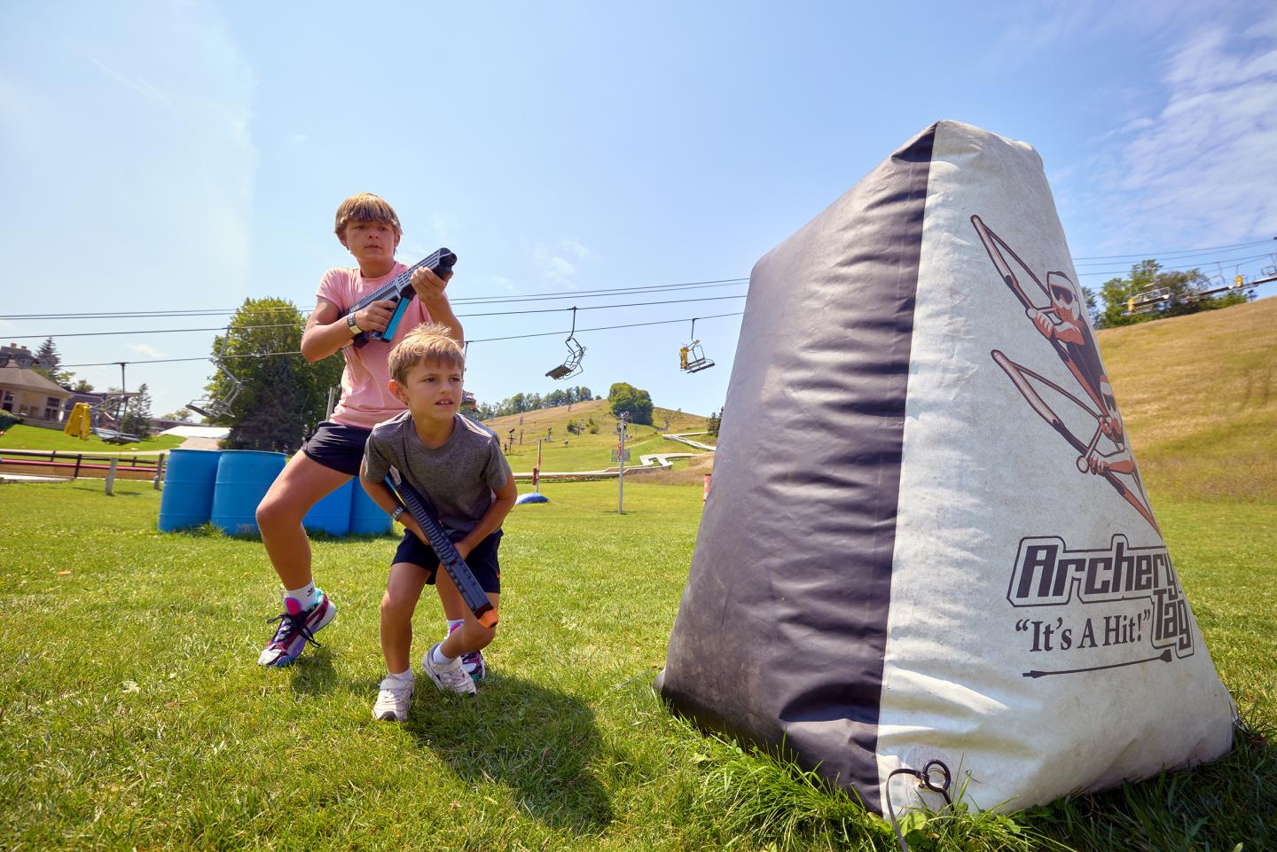 Two kids with toy blasters crouch behind a triangular inflatable on a grassy field.