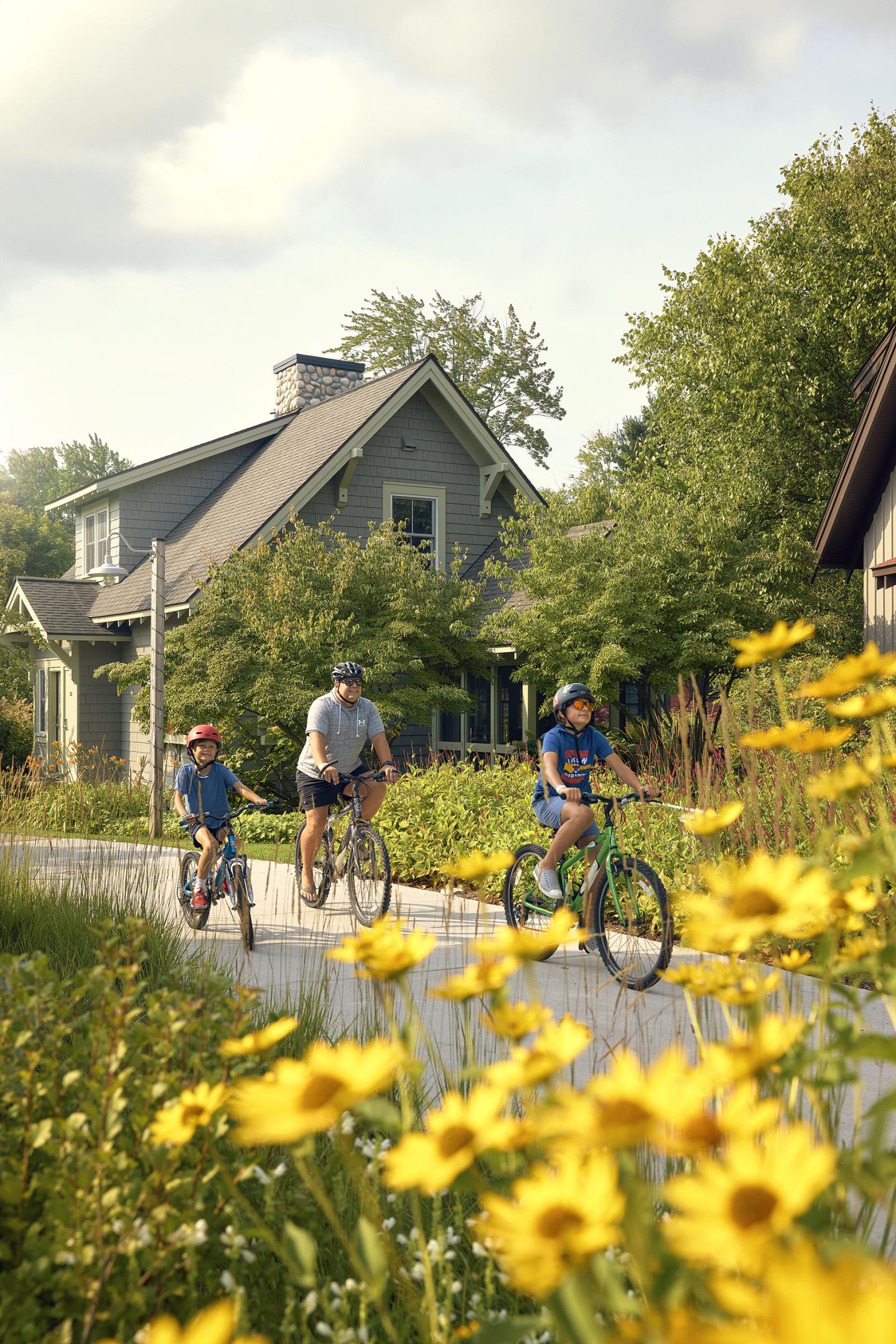 Family riding bikes on a path by yellow flowers and houses.